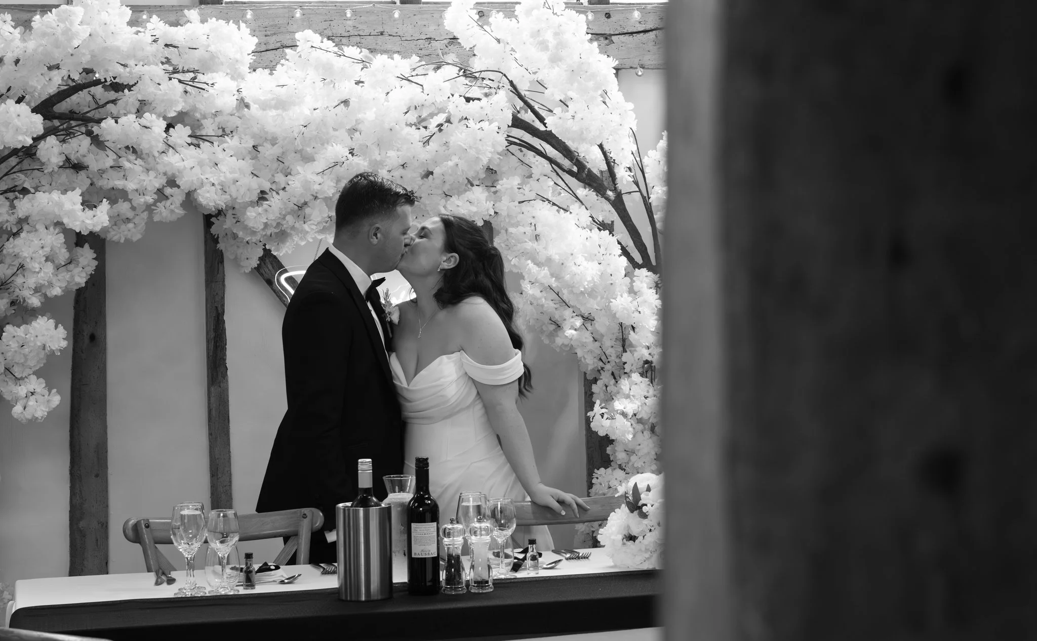 A black and white photo of a bride and groom sharing a kiss at their wedding reception, standing by a decorated table with wine bottles and glasses, and a floral backdrop. Anne Wills Photography Maidens barn Essex