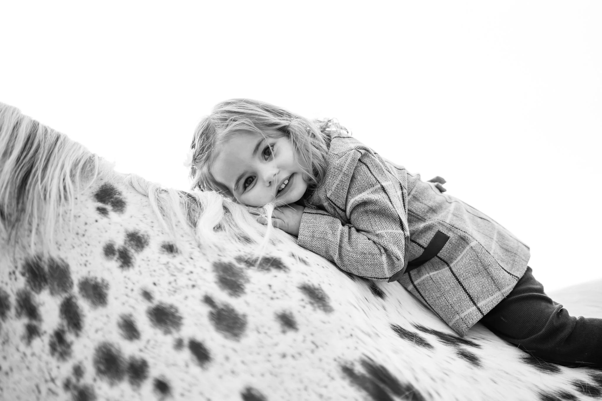 A young girl with curly hair laying her head on a spotted horse's neck, smiling in an outdoor setting. photographed in felsted essex. Anne Wills Photography 