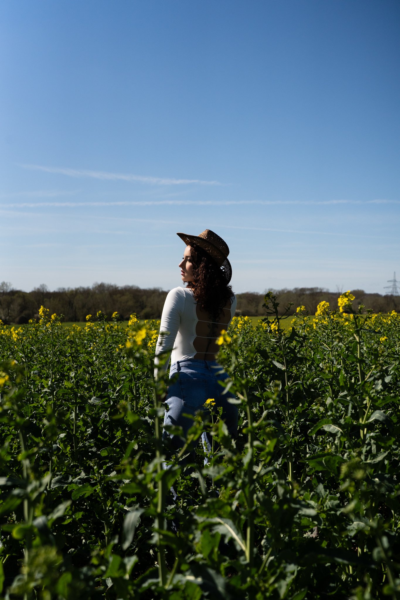 A woman stands in a green field of plants with yellow flowers, wearing a straw hat and a white backless top, under a blue sky with wispy clouds. photographed in coggehall essex. Anne Wills Photography 