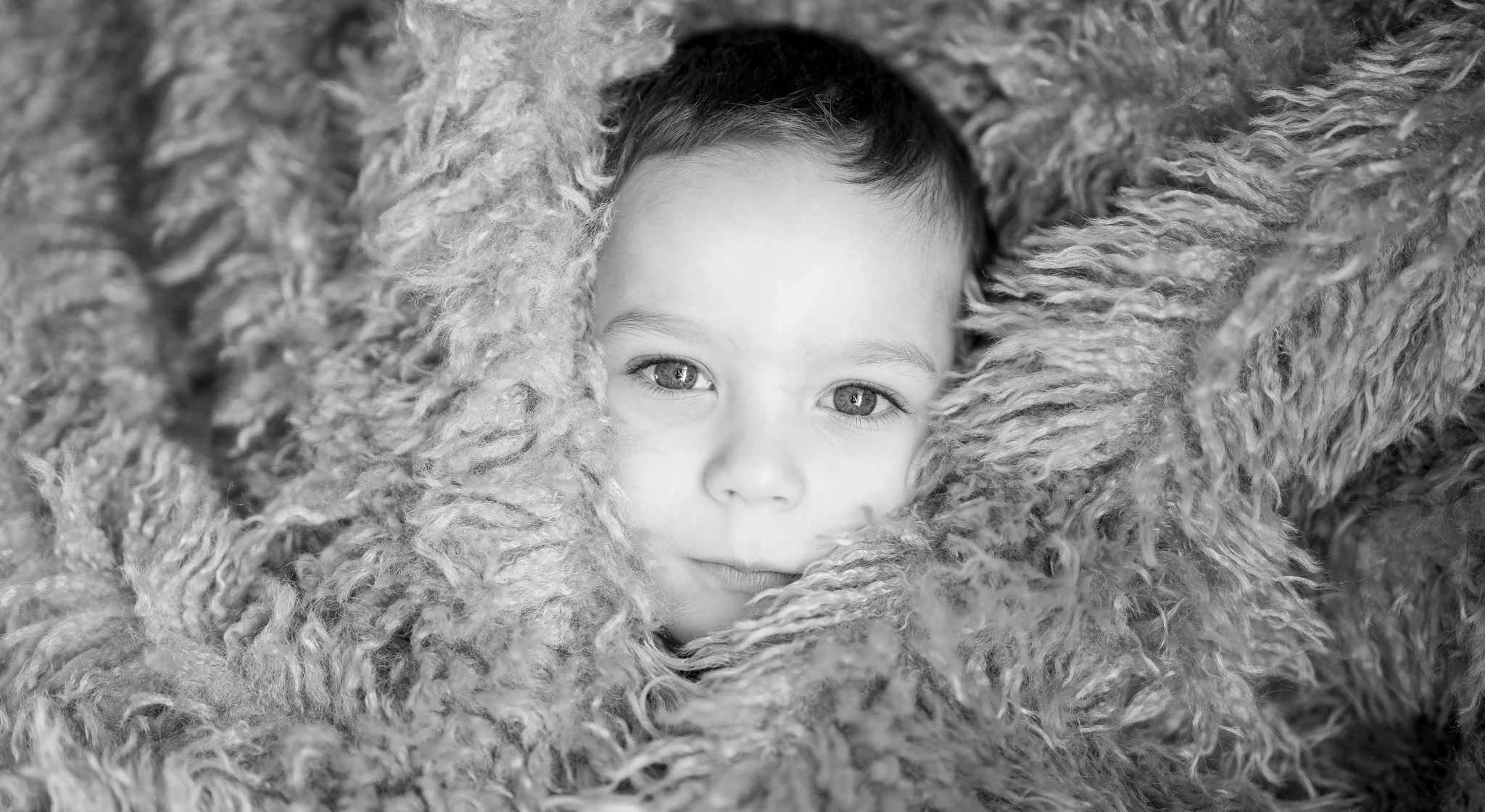 A young child with large, bright eyes gazes out from amidst fluffy, textured material, creating a cozy, intimate scene. photographed in Braintree, Essex. Anne Wills Photography 