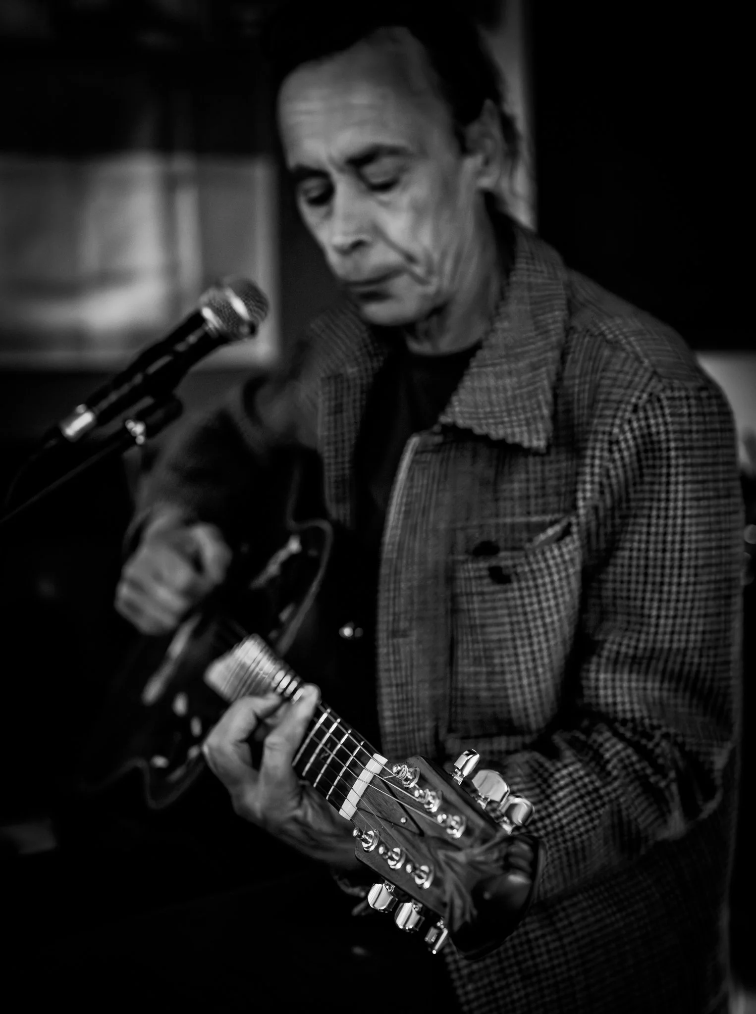 Black and white photo of a man with a checkered blazer playing an electric guitar with a microphone nearby.