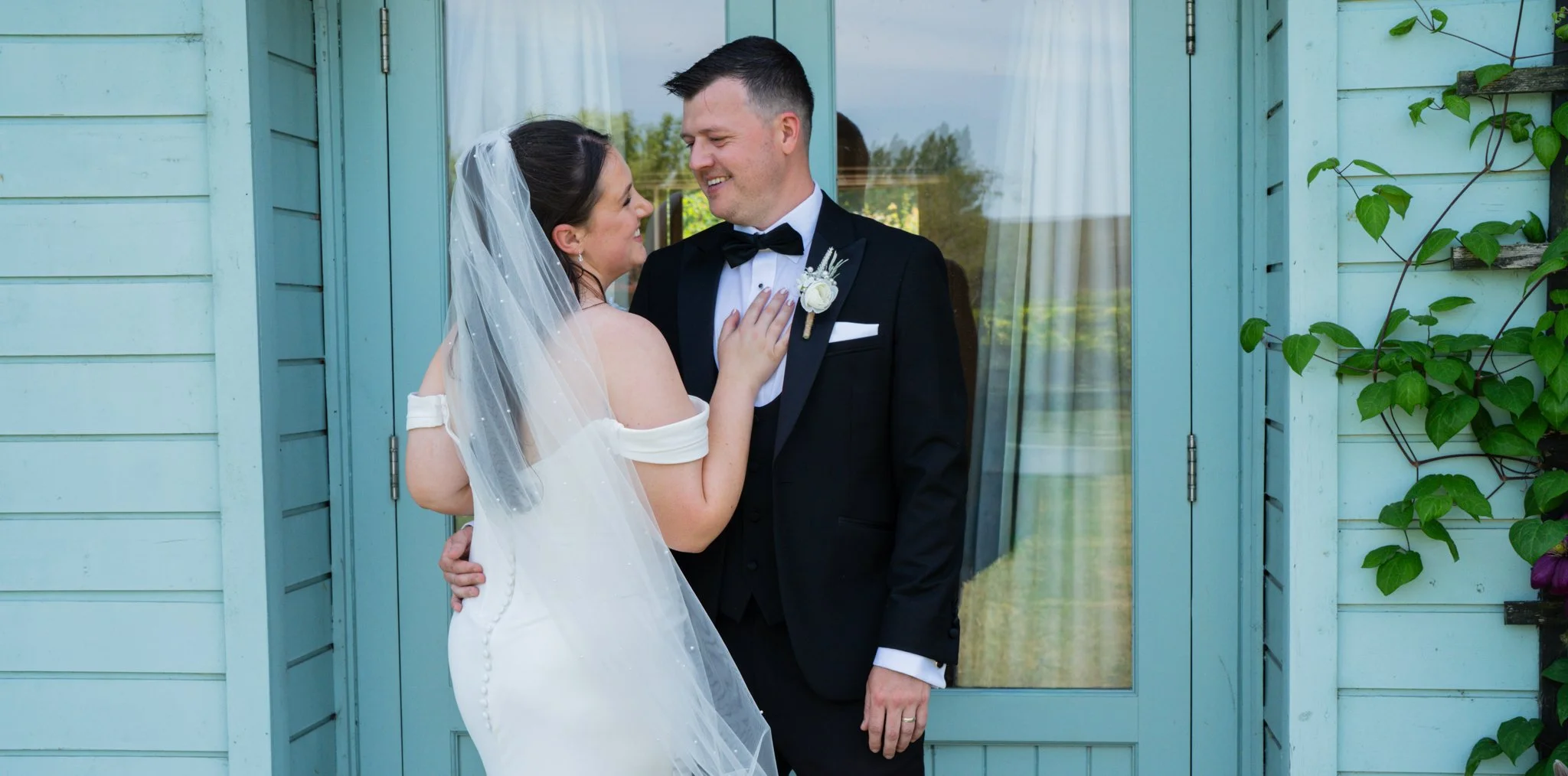 A newlywed couple in wedding attire smiling and gazing at each other outside a light blue house with a glass door. Anne Wills Photography Maidens barn Essex