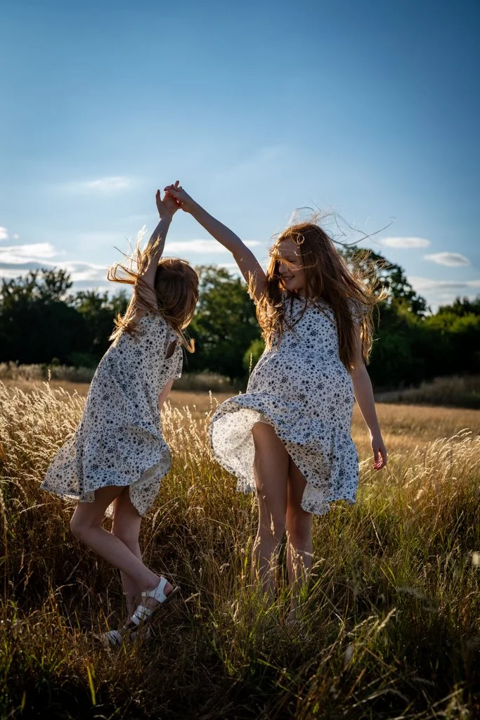 Two young girls in matching floral dresses playing and dancing in a field of tall grass during sunset.