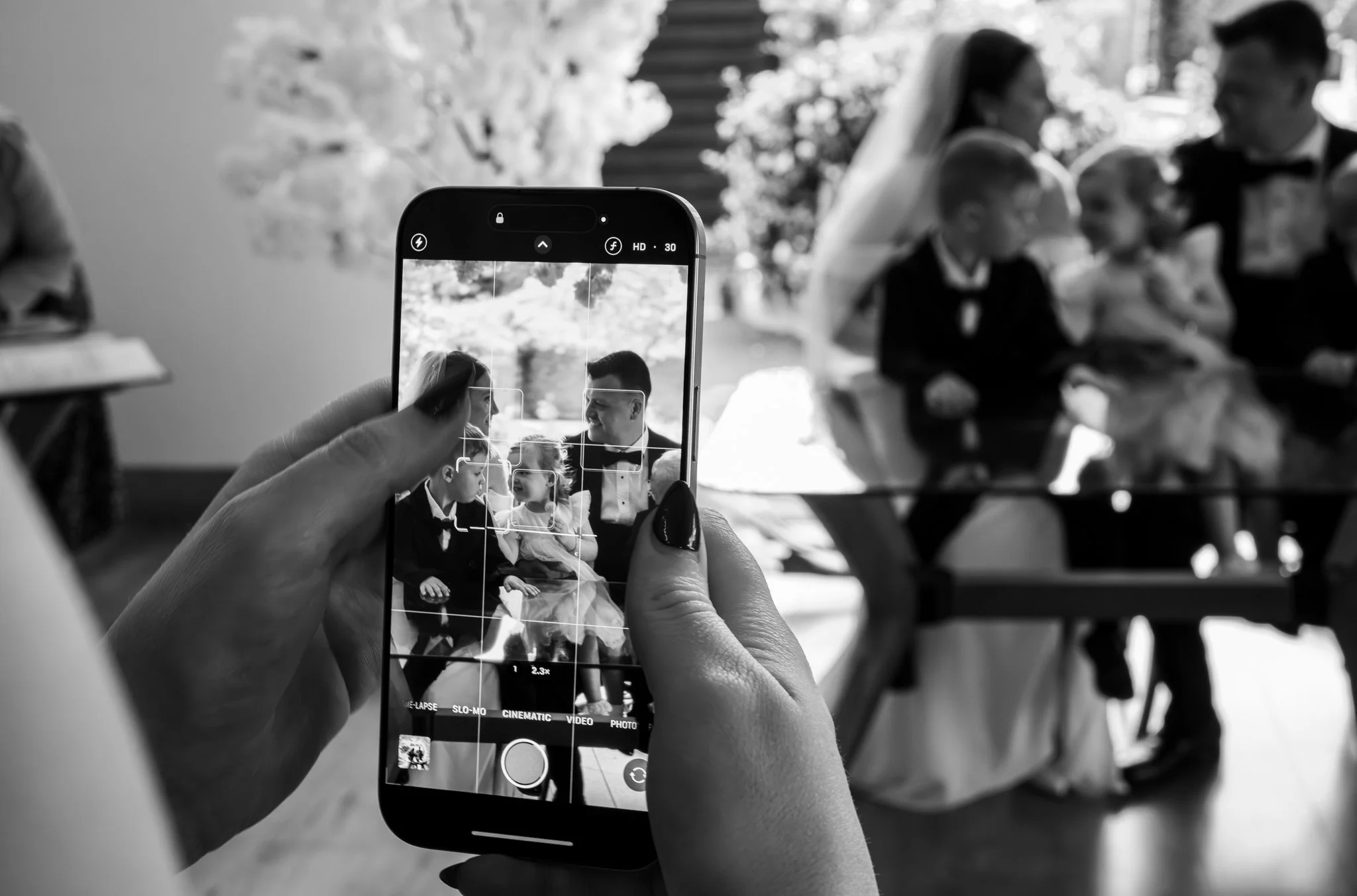 A person is taking a photo of a family at an event with a smartphone. The family, visible on the phone's screen, includes two children wearing tuxedos and a woman in a dress, with a man in a tuxedo. The background is slightly blurred, showing trees a