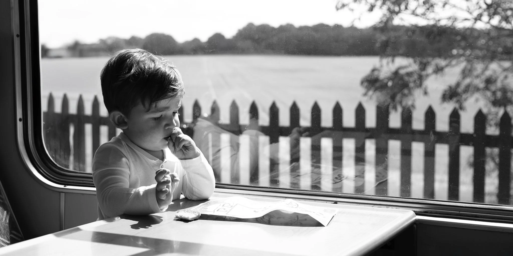 A young boy sitting at a table, looking out a window on a bus or train, with a fence and trees visible outside. Anne Wills Photography 