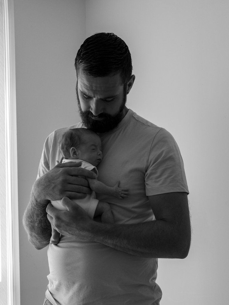 A man with a beard holding a newborn baby close to his chest, looking down at the baby in a gentle and caring manner.