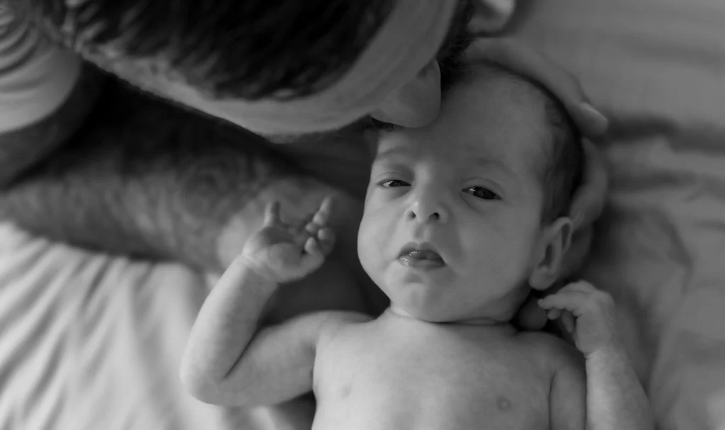 A newborn baby with a chubby face, lying on a bed and looking at the camera, while an adult leans over, gently touching the baby's head.