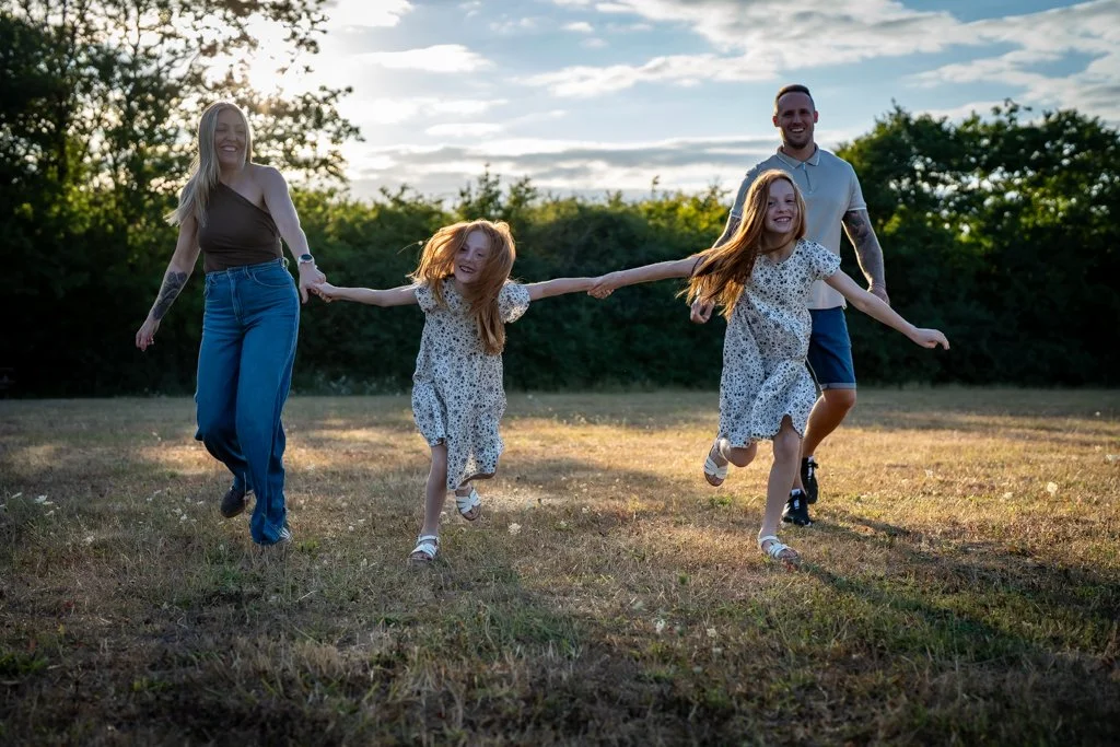 A family of four running outdoors in a grassy field at sunset, holding hands and smiling.