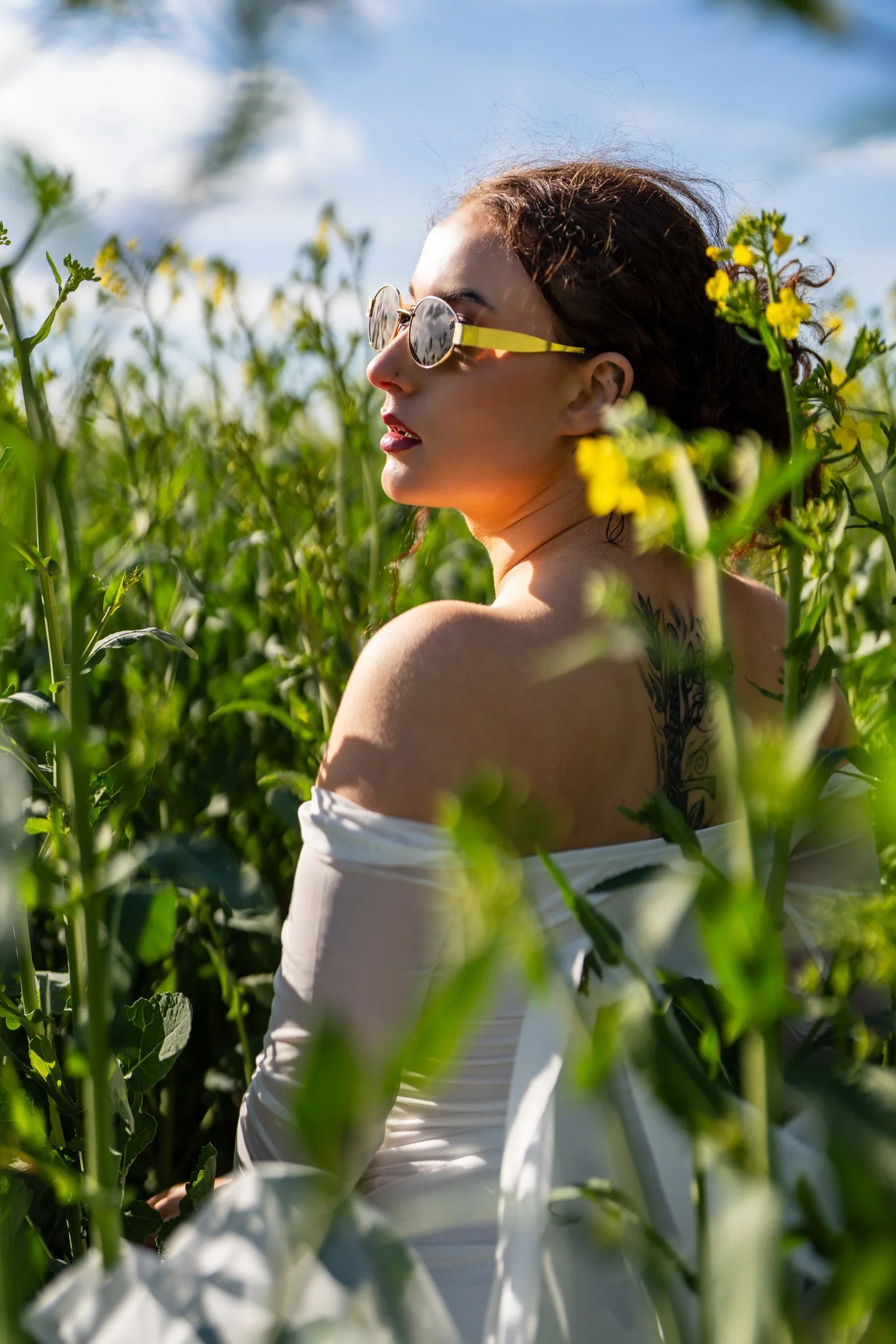 A woman with dark curly hair, wearing sunglasses and a white off-the-shoulder top, is standing in a field of yellow flowers during daytime, facing sideways. photographed in a field in coggeshall, Essex. Anne Wills Photography 