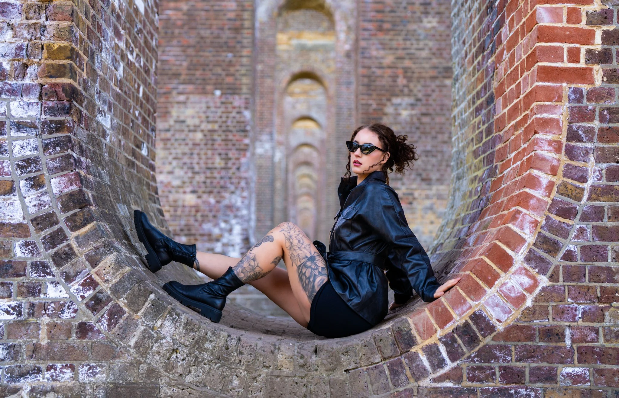 A woman with tattoos wearing black boots, black leather jacket, and sunglasses sitting inside a brick tunnel or archway. photographed in colchester essex Anne Wills Photography 