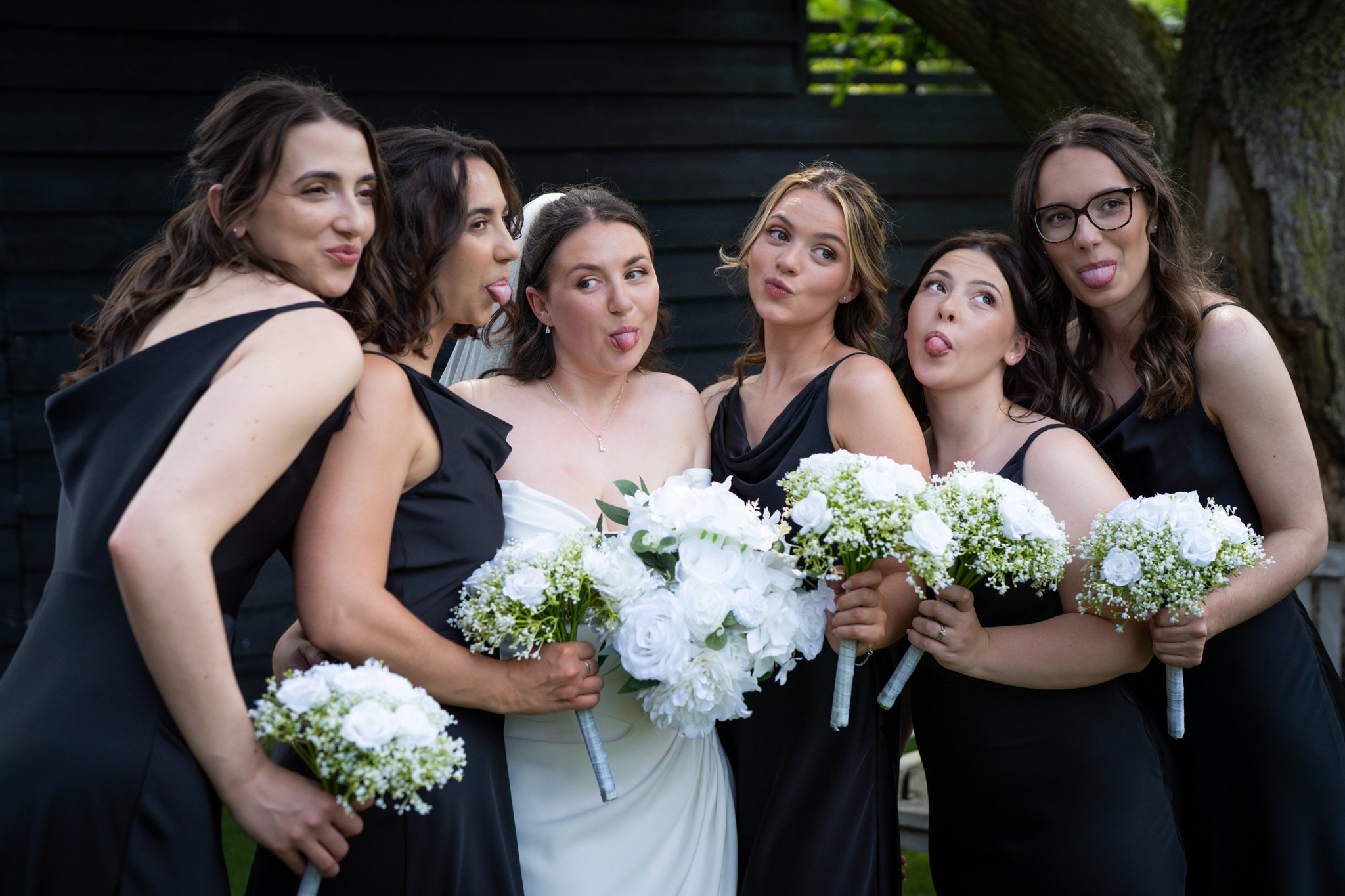 A bride and her five bridesmaids making silly faces, sticking out their tongues, while holding bouquets of white flowers outdoors during a wedding. Anne Wills Photography Maidens barn Essex