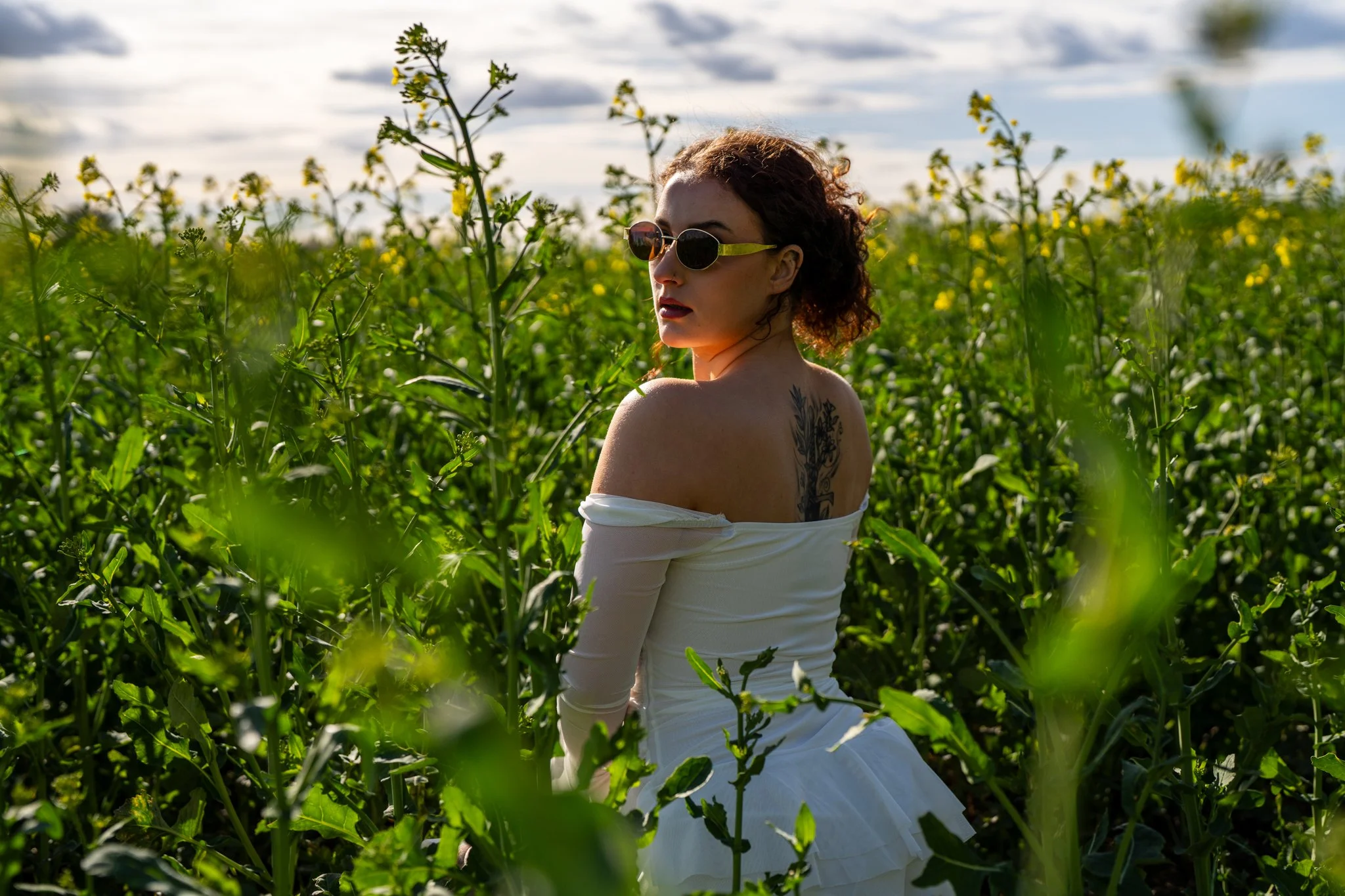 A woman with curly hair wearing sunglasses and a white off-the-shoulder dress sitting in a green field of tall plants with yellow flowers under a partly cloudy sky.