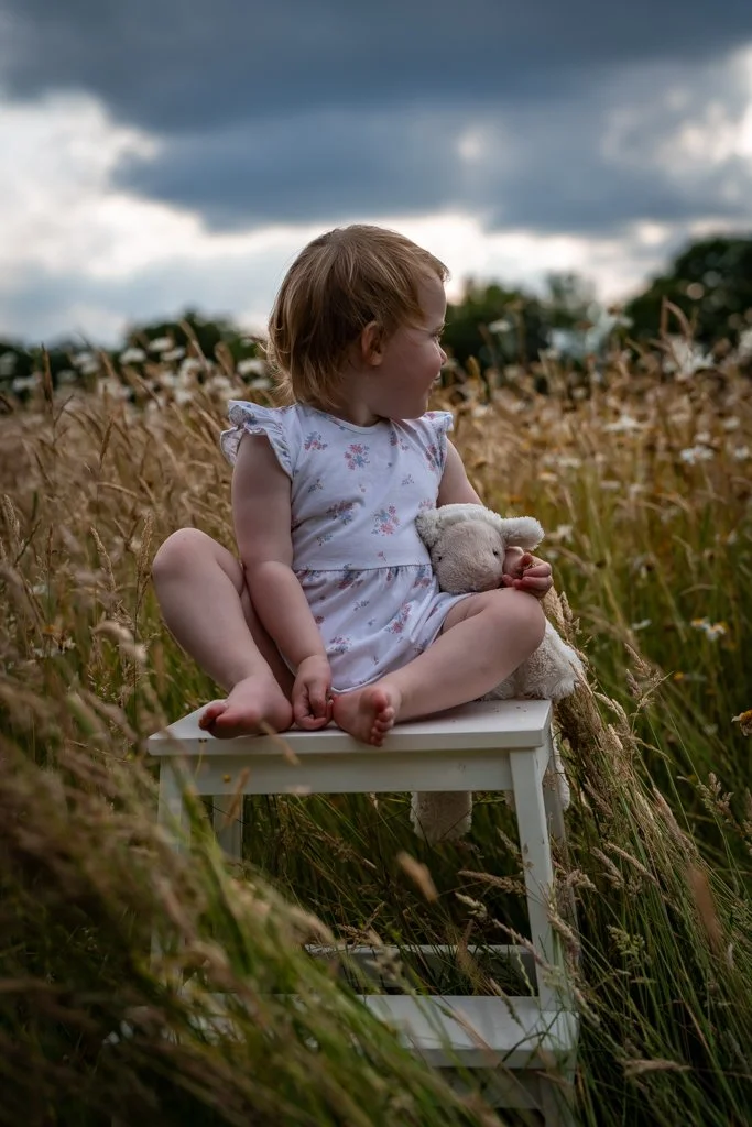A young girl sitting cross-legged on a small white chair in a field of tall grasses holding a stuffed animal, with dark clouds in the sky. Anne Wills Photography. Braintree, Essex