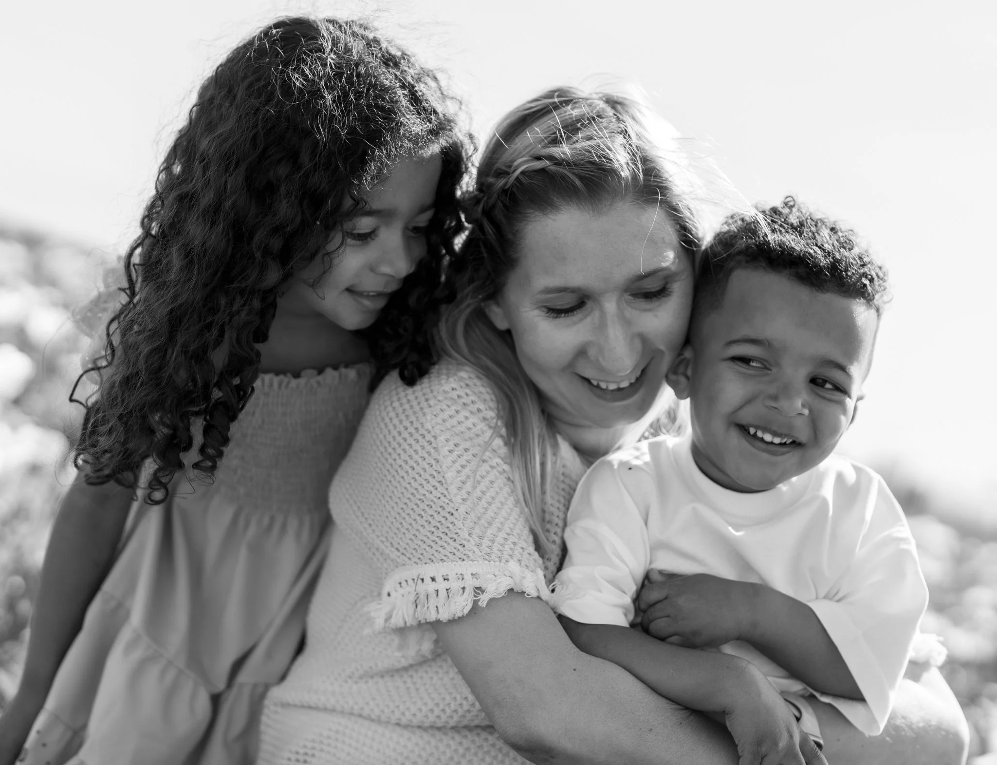 Four people, three children and one adult, smiling and hugging each other outdoors in sunlight, in a black and white photograph. hatters farm tulip festival, Essex. Anne Wills Photography 
