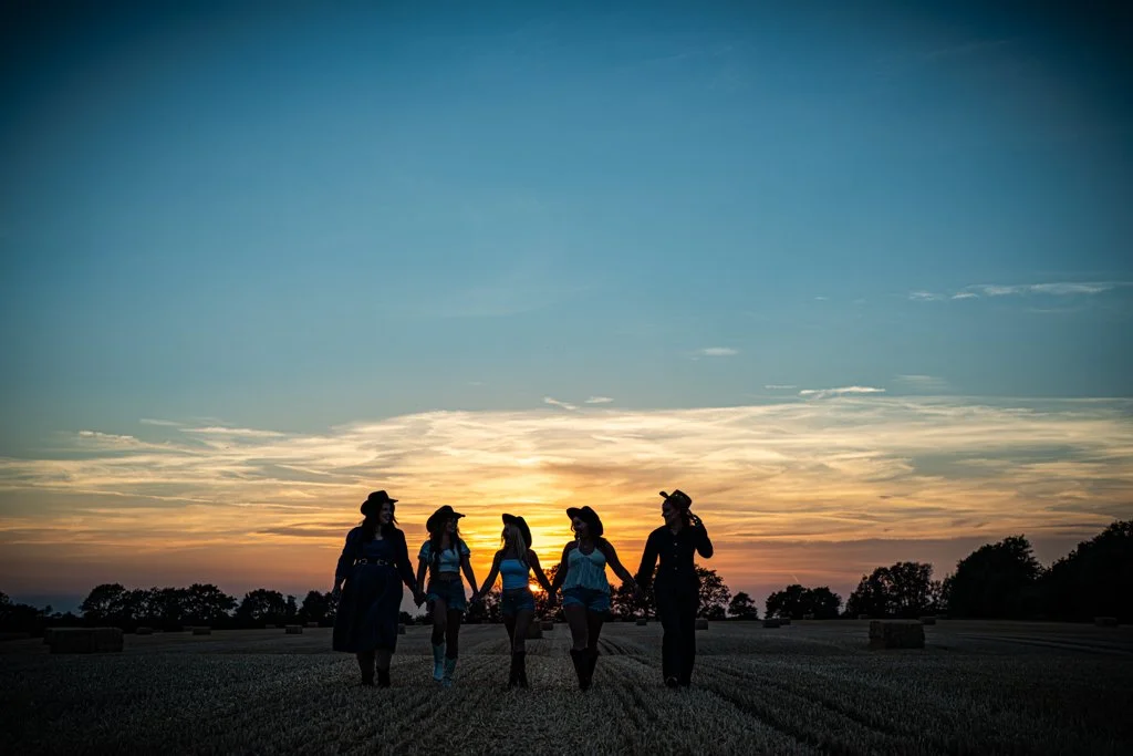 Group of women in cowboy hats holding hands walking through a field at sunset.