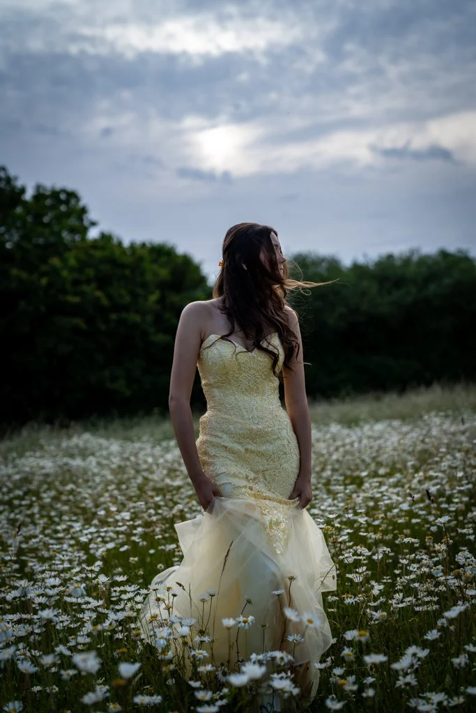 A woman in a yellow strapless dress standing in a field of white flowers during cloudy weather. Anne Wills Photography. Braintree, Essex