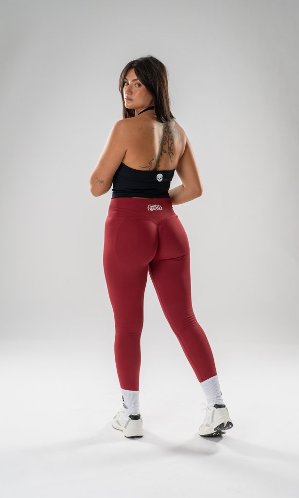 Woman wearing black sports top, red leggings, white athletic shoes, and white socks, standing in a studio with a plain gray background.