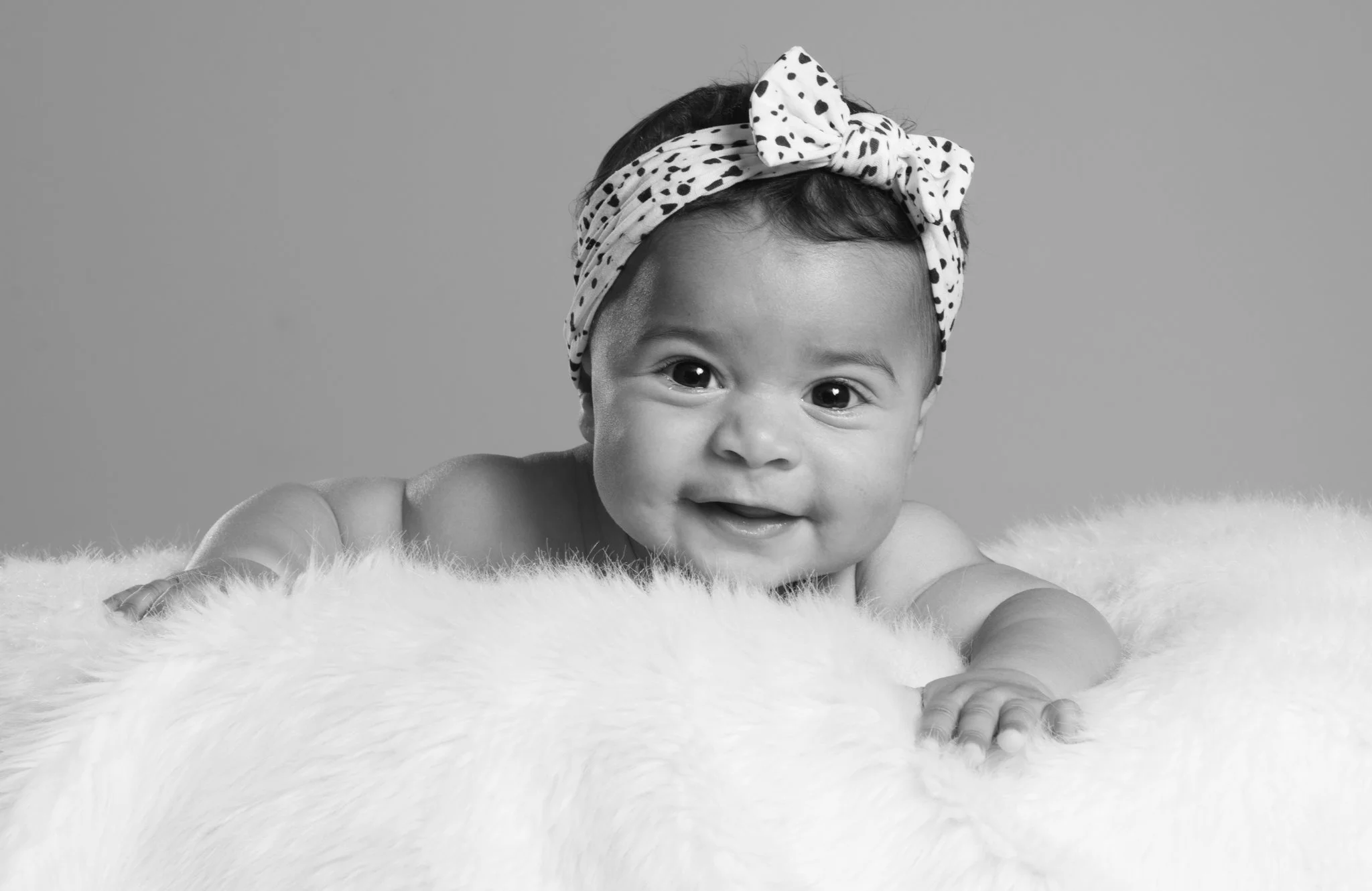 A smiling baby girl lying on a soft, fluffy surface, wearing a polka dot headband and looking directly at the camera. photographed in Braintree, Essex. Anne Wills Photography 