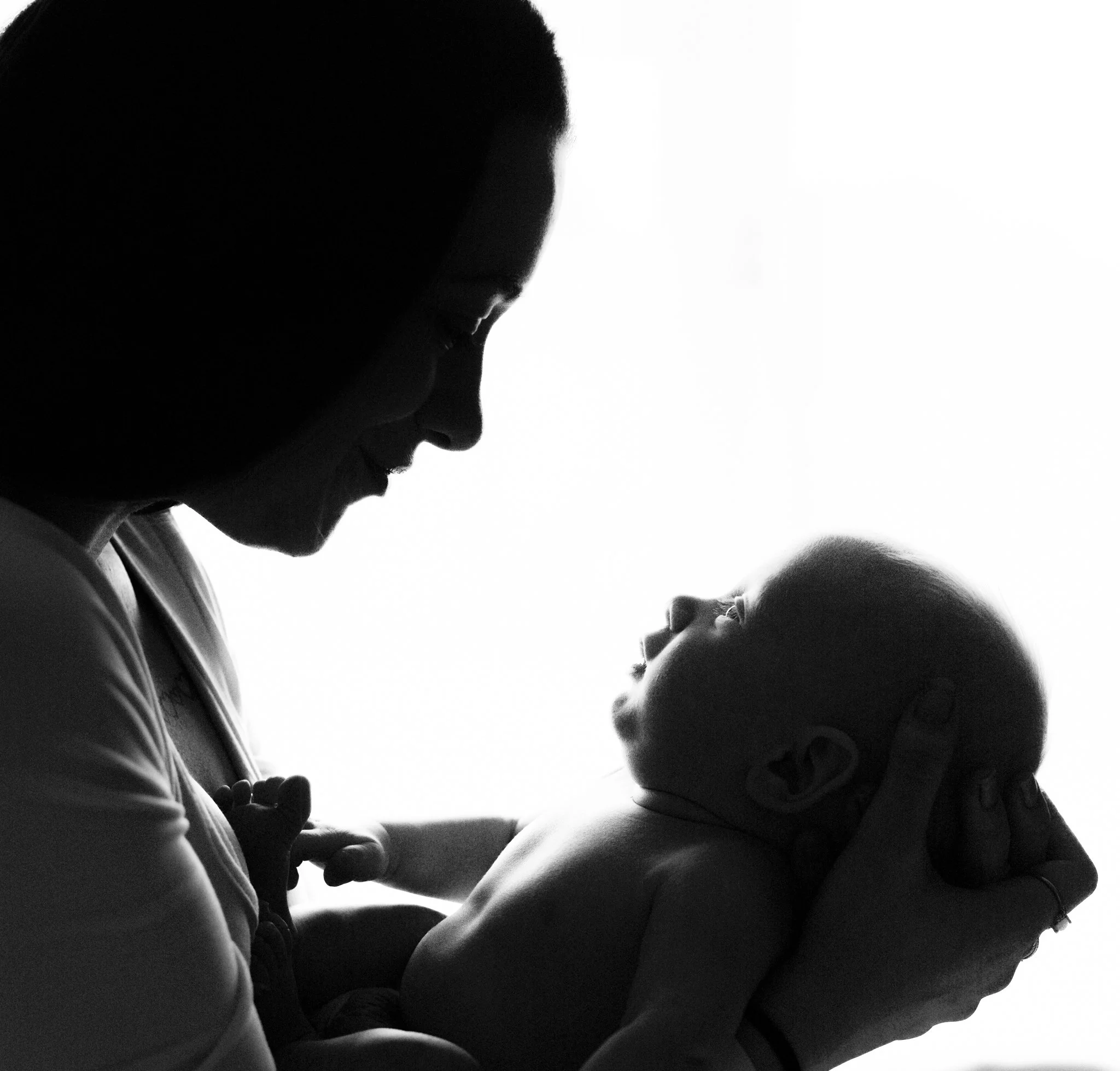 A black and white photo of a woman and a child facing each other and holding hands, with their heads bent towards each other. Photographed at customers home in colchester, Essex. Anne Wills Photography 