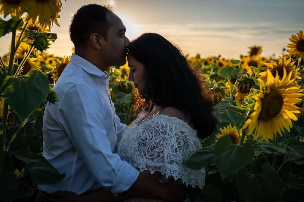A couple standing close together in a sunflower field at sunset, with the man kissing the woman's forehead.