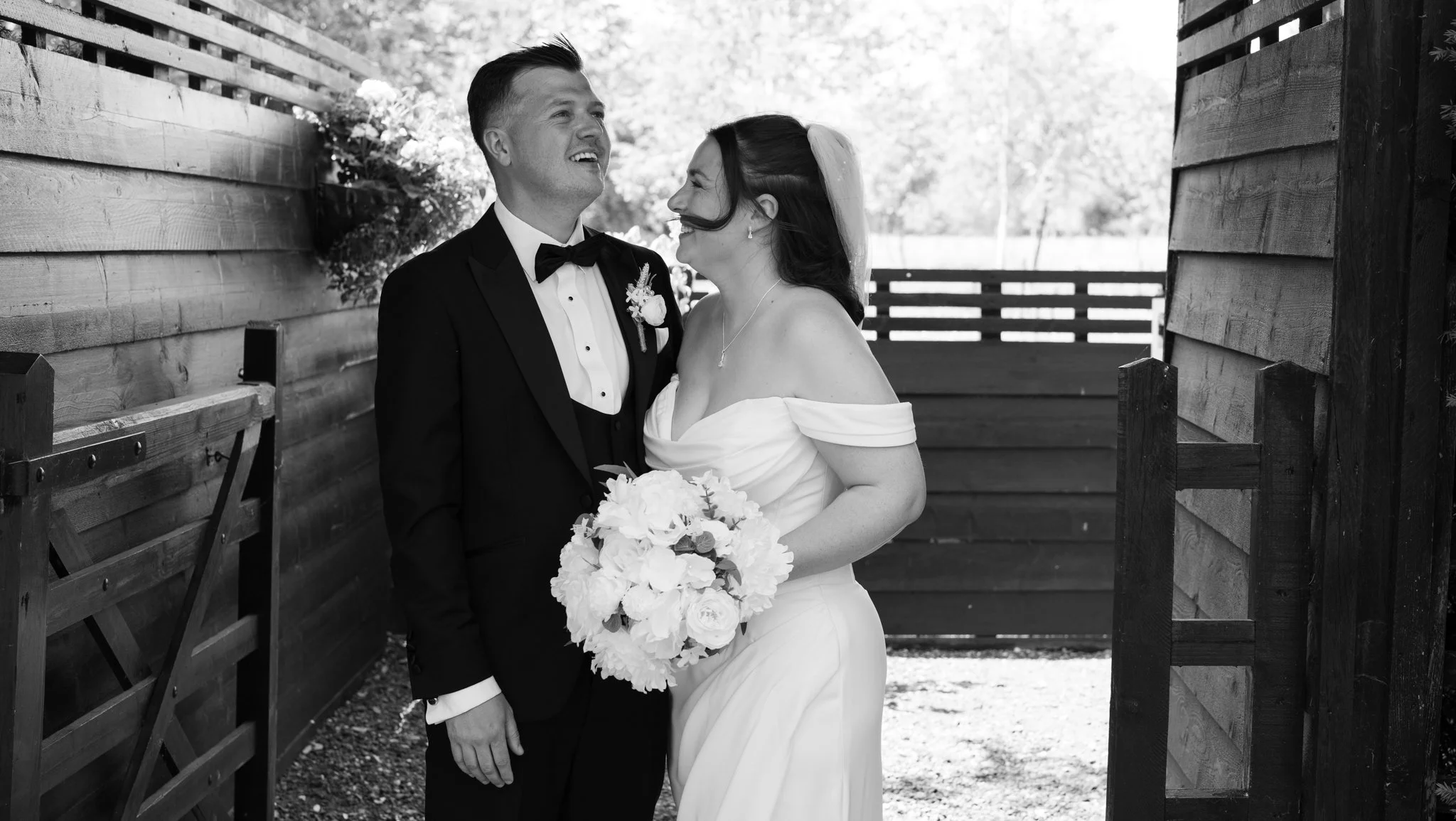 Black and white photo of a bride and groom smiling and looking at each other outdoors, with the bride holding a bouquet of flowers.  Anne Wills Photography Maidens barn Essex