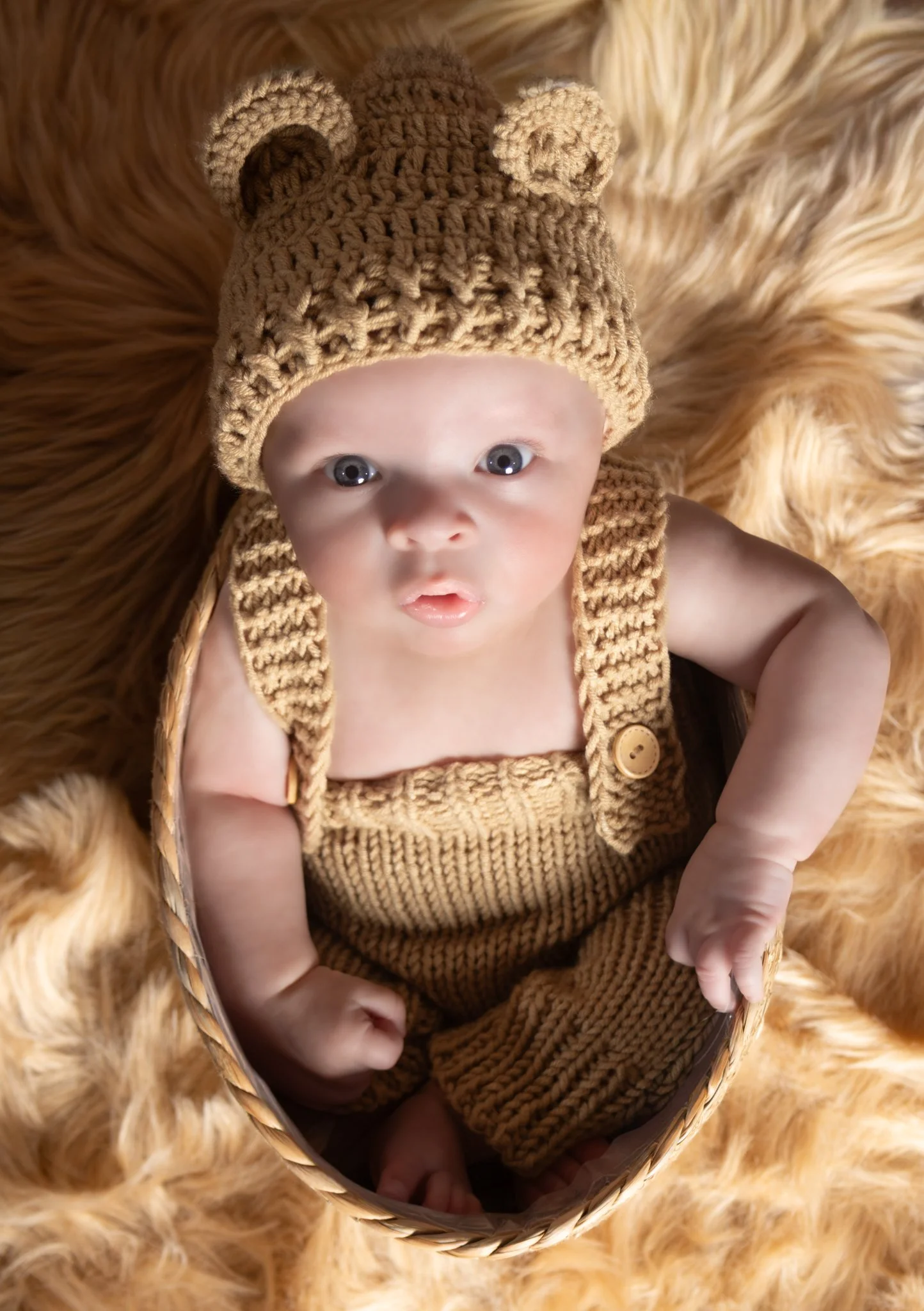 Close-up of a baby dressed in brown knitted outfit with a bear-themed hat, lying in a basket on a faux fur blanket.