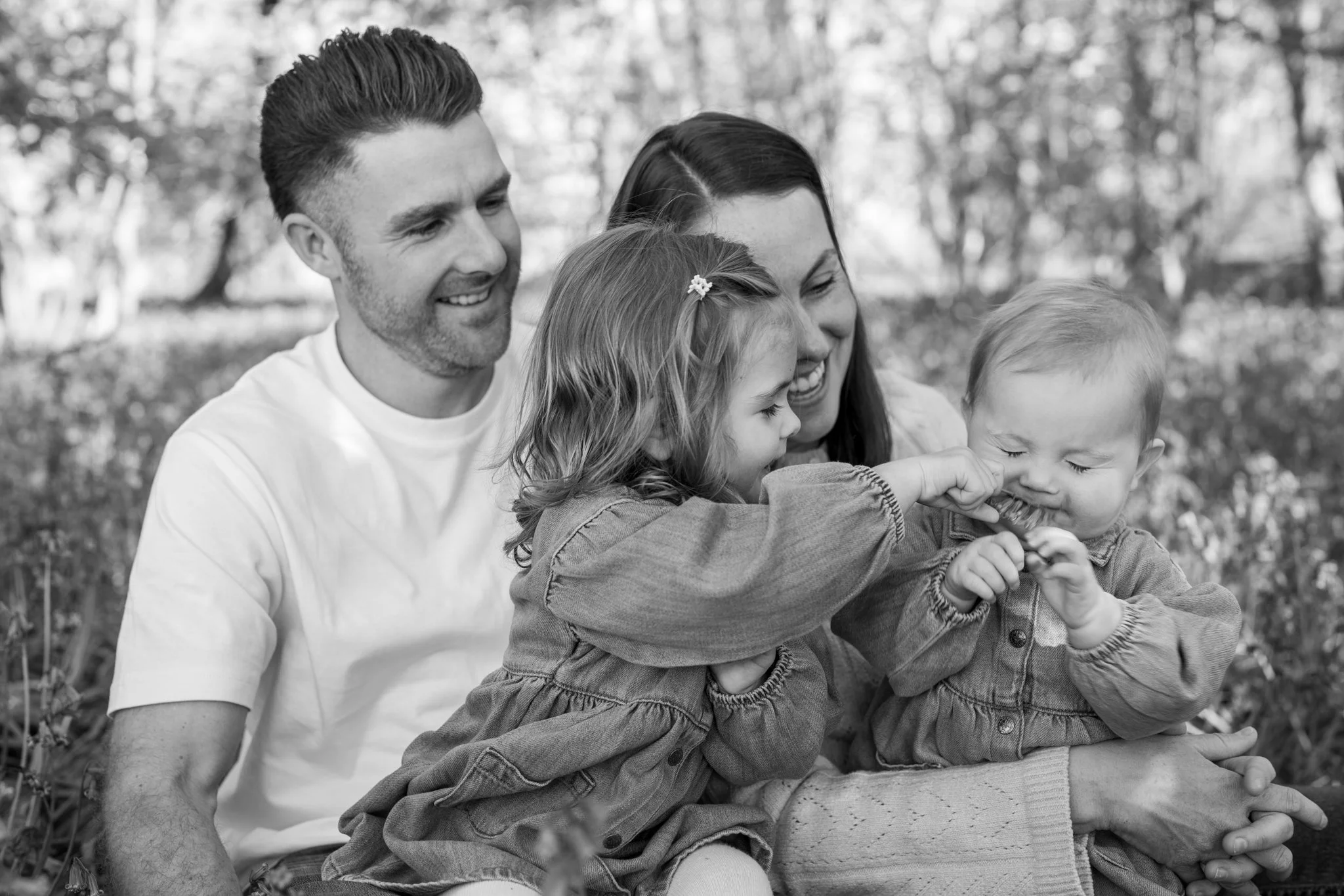 A smiling family of four outdoors, sitting on a blanket, with trees in the background. The parents are a man and a woman, and they are playing with their two children, a girl and a boy, who are sitting in their laps. Salford, Essex. Anne Wills Photog