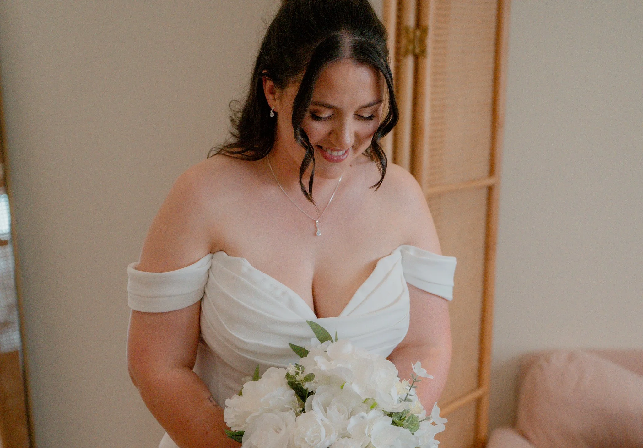 A woman wearing an off-the-shoulder white wedding dress, holding a bouquet of white flowers, and smiling with her eyes closed. Anne Wills Photography Maidens barn Essex