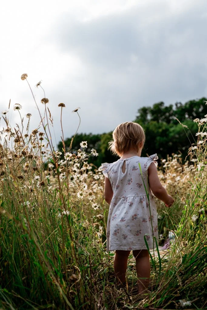 A young girl in a white floral dress walking through a field of tall wildflowers, with trees and a cloudy sky in the background. Anne Wills Photography. Braintree, Essex