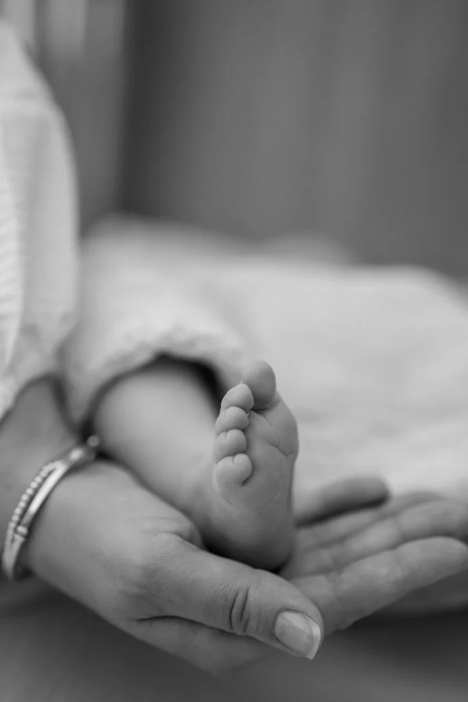 A small baby's foot resting on an adult's hand, focusing on the baby's tiny toes with a blurred background.