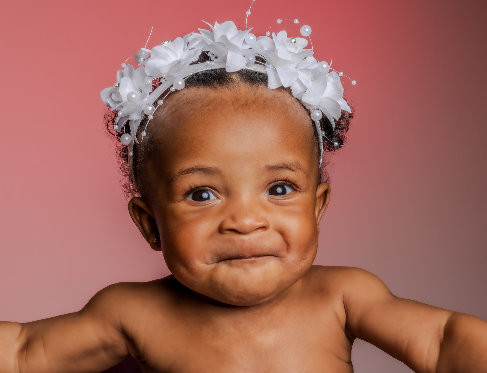 A young child with blue eyes and short curly hair wearing a white decorative headband with flowers and pearls, posing with a playful facial expression against a pink background. Photographed in Studio in Essex. Anne Wills Photography 