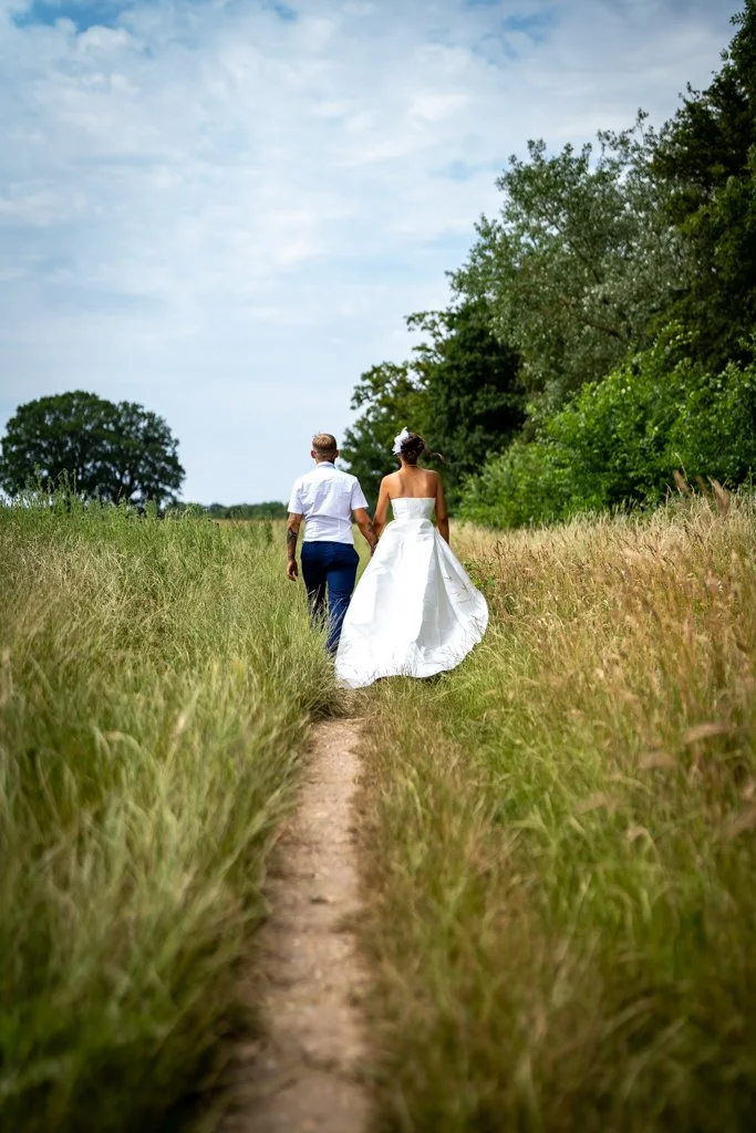 A bride and groom walking hand in hand along a narrow dirt path through tall grass, with trees and cloudy sky in the background. Anne Wills Photography. Braintree town hall, Essex