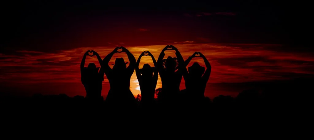 Silhouettes of five people standing in a line with their arms raised upwards making heart shapes with their hands, during a vibrant sunset with a reddish-orange sky.