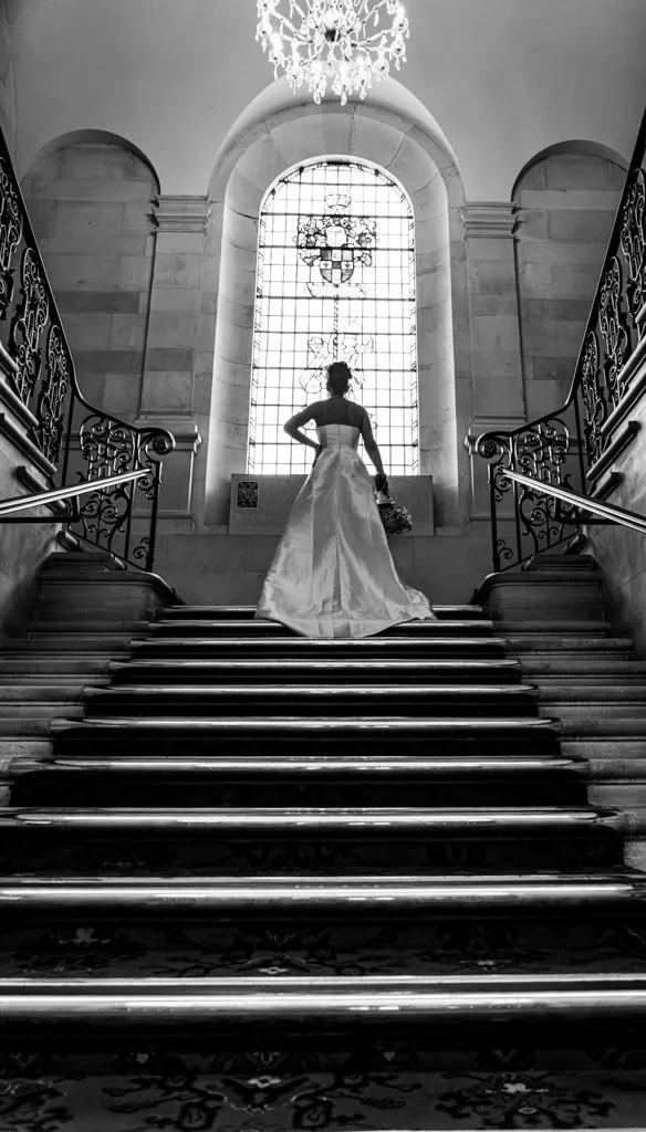 A bride standing at the top of a grand staircase inside a church, facing a large stained glass window with her back to the camera, holding a bouquet of flowers.
anne wills photography . Braintree, Essex