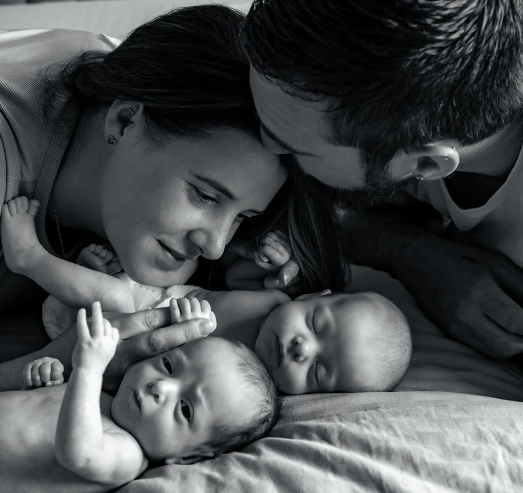 Black and white photo of a couple lying down with two babies, one with eyes open and one sleeping, as the woman and man gently touch and kiss the babies.