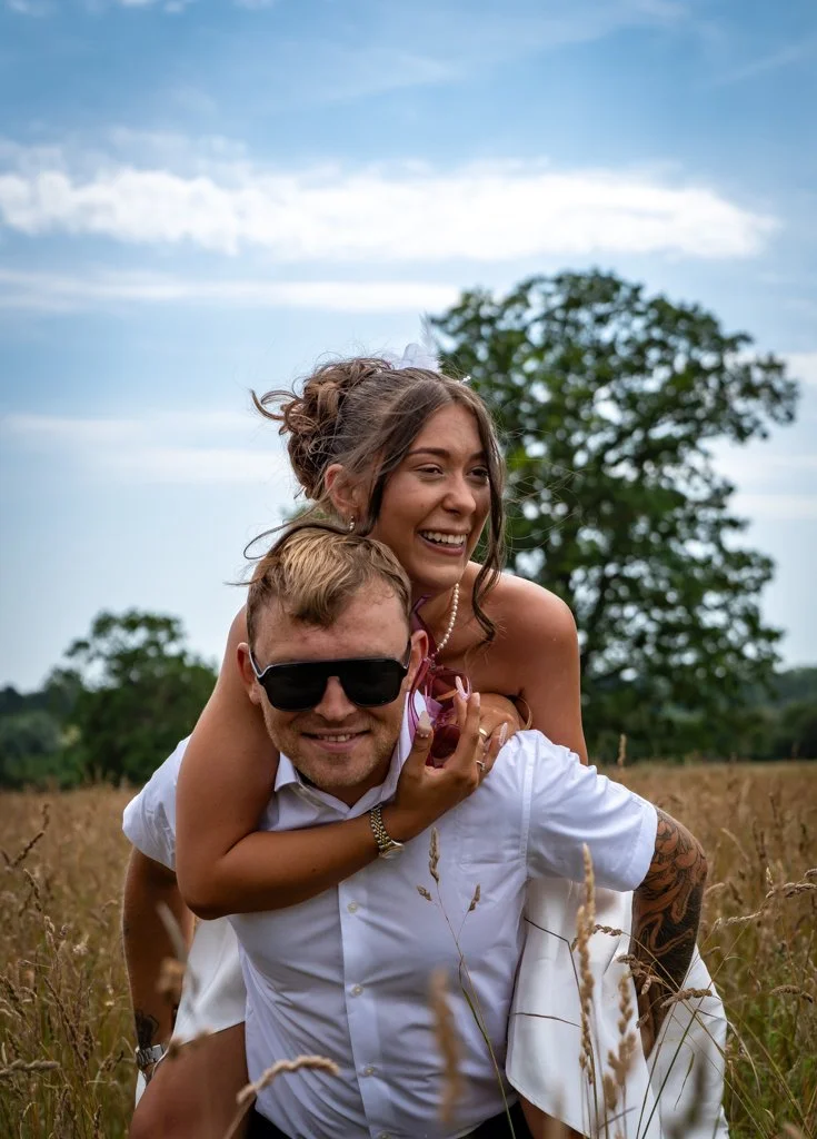 A smiling woman giving a piggyback ride on a man with sunglasses in a field of tall grass, with a large tree and cloudy sky in the background. Anne Wills Photography. Braintree town hall, Essex