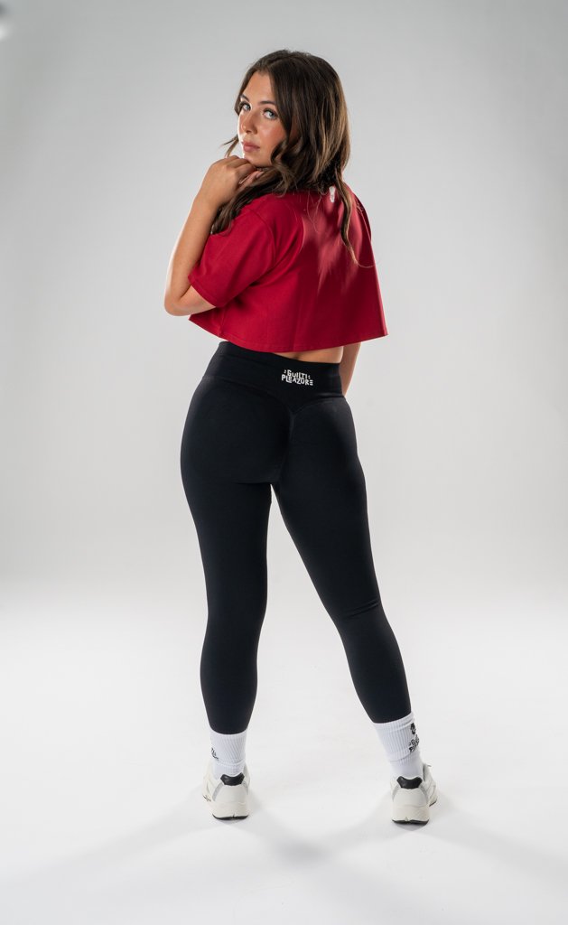Young woman in a red cropped t-shirt, black leggings, white socks, and sneakers standing with her back to the camera, head turned to the side, on a plain gray background.