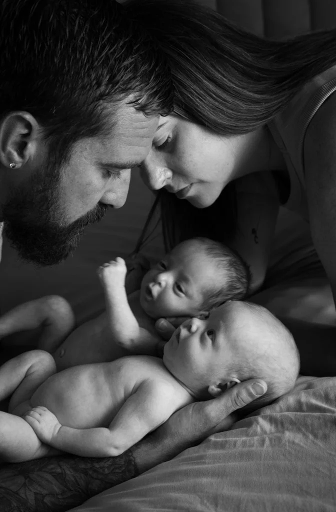 A black and white photo of a family with two infants. The mother and father are leaning overhead, with their foreheads touching, looking lovingly at each other and the babies. The father is holding one baby, and the mother is tenderly holding the oth