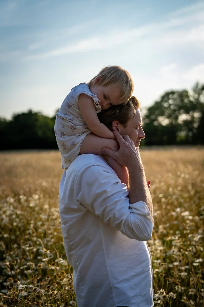 A man carrying a young girl on his shoulders in a field during sunset. Anne Wills Photography. Braintree, Essex