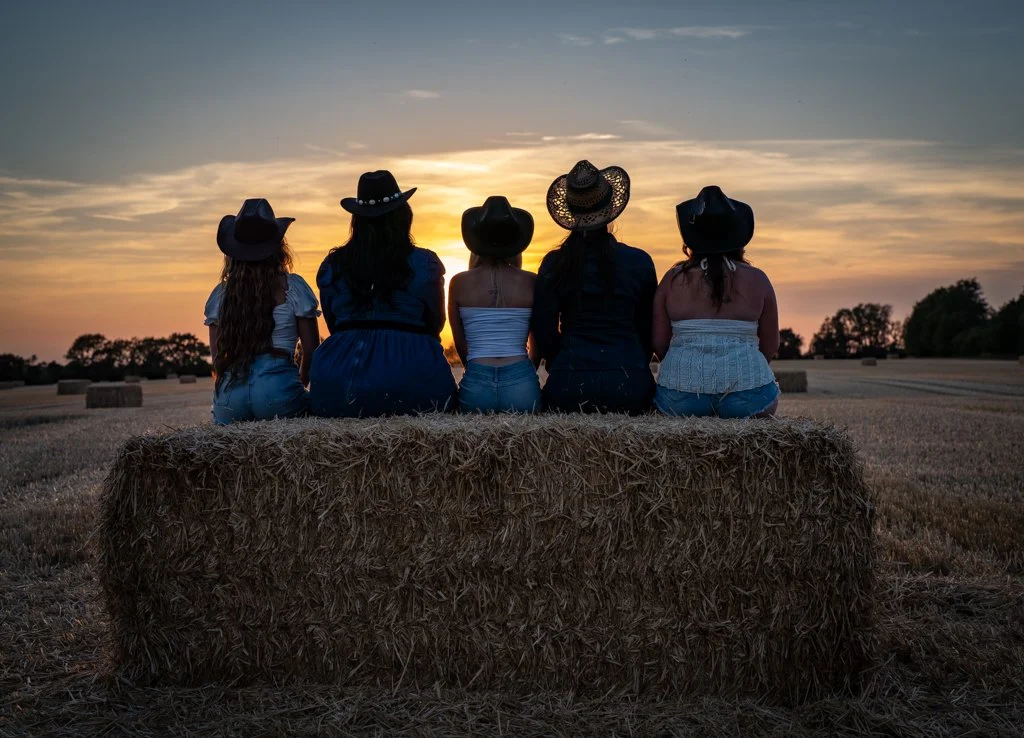 Five women wearing cowboy hats sitting together on a hay bale in a field during sunset.