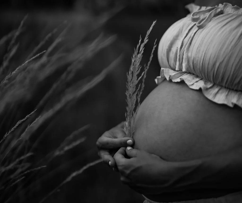 Close-up of a pregnant woman's belly with one hand holding a dried grass stem, outdoors, in black and white.