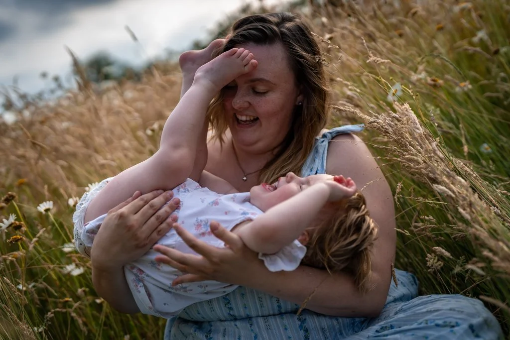 A woman and a young girl enjoying a playful moment in a grassy field with flowers, smiling and laughing together. Anne Wills Photography. Braintree, Essex