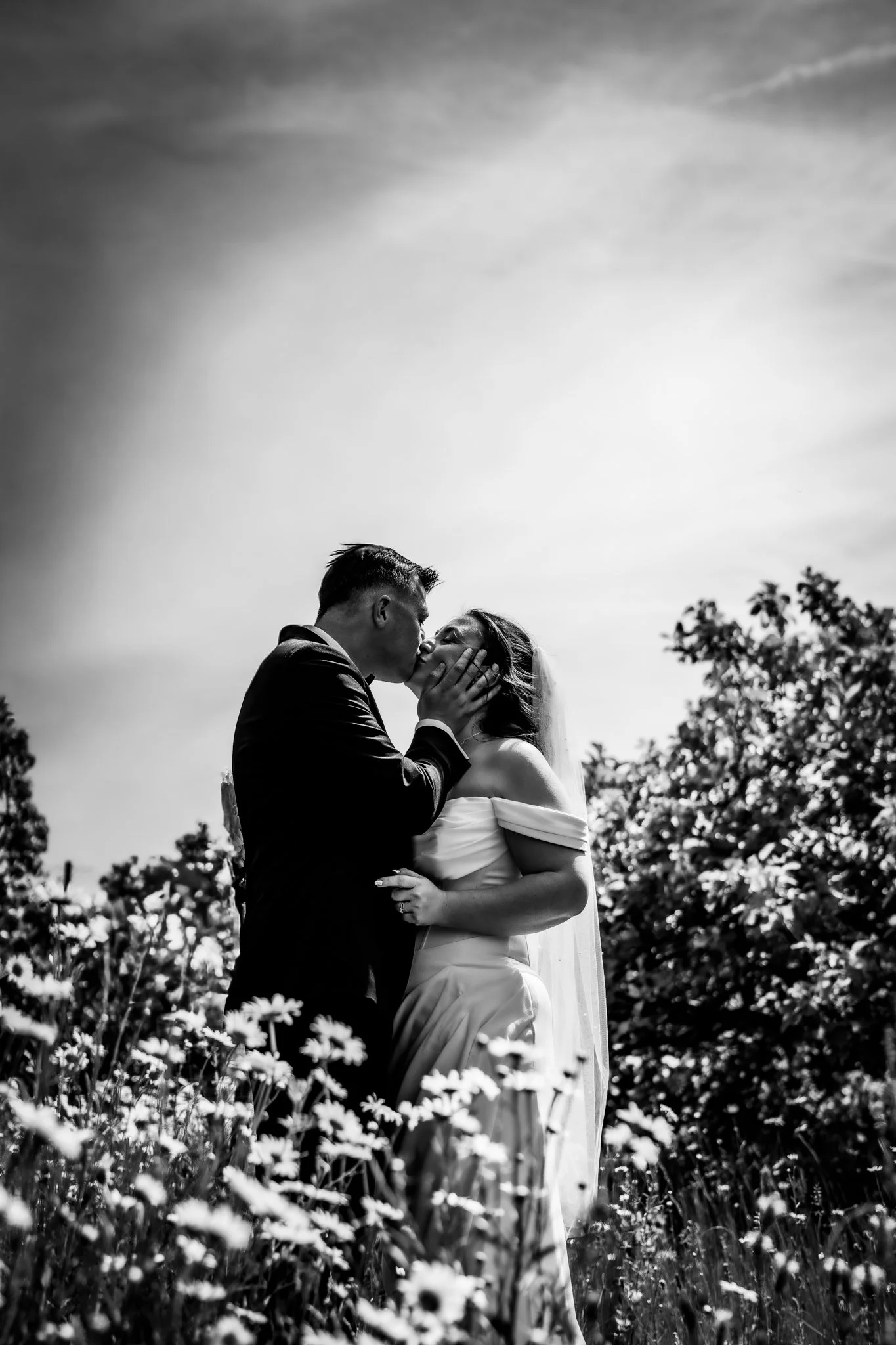 A black and white photo of a couple in wedding attire sharing a kiss outdoors amidst flowers and trees, with a partly cloudy sky in the background.