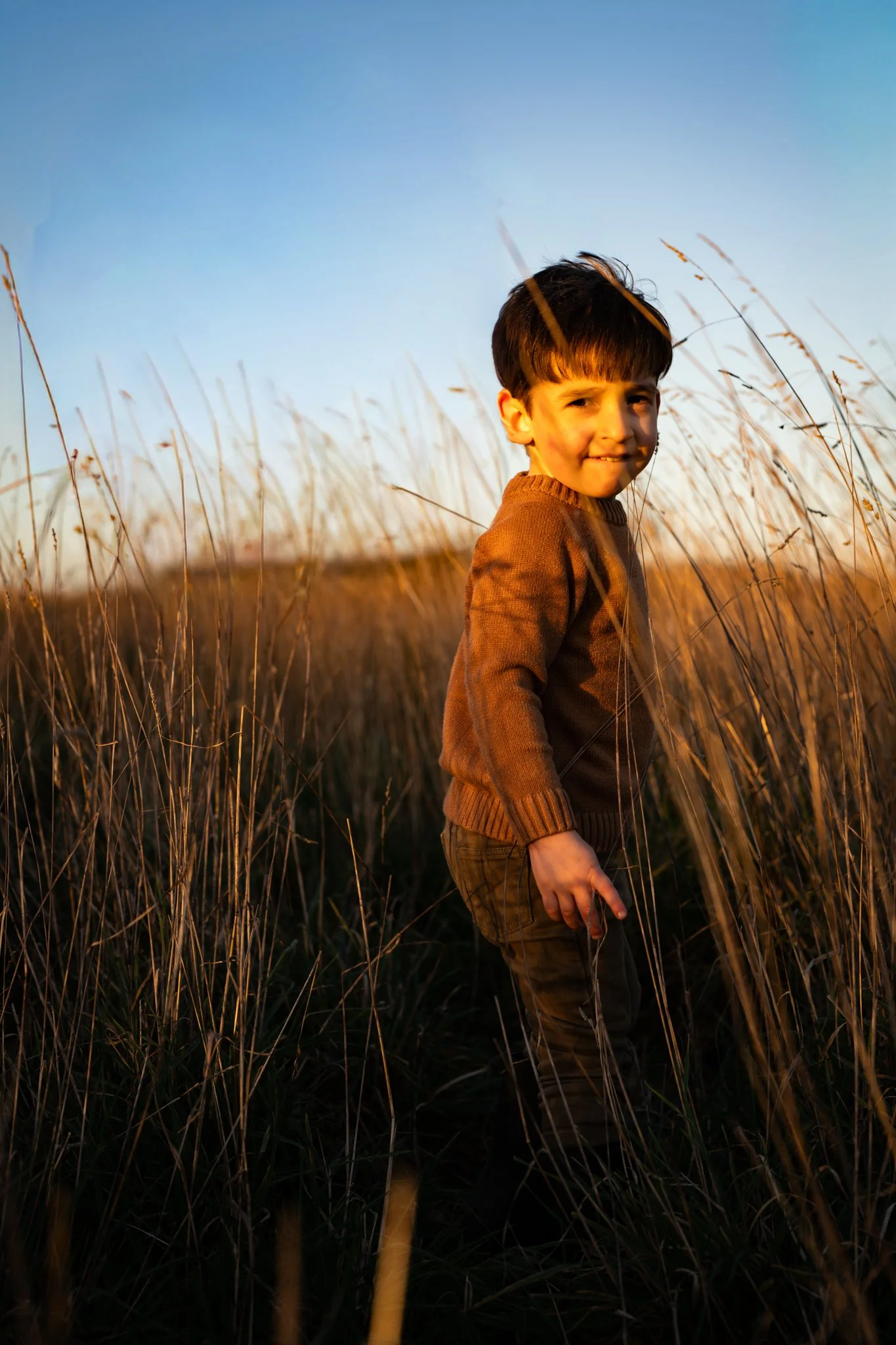 A young boy standing in a field of tall grass during sunset, smiling and looking back at the camera. mocking, Essex. Anne Wills Photography 