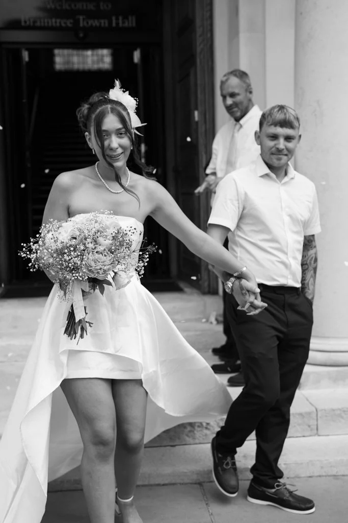 A woman in a strapless wedding dress holding a bouquet of flowers and a man in a white shirt and dark pants holding hands, with another man in the background outside a building. Anne Wills Photography. Braintree town hall, Essex