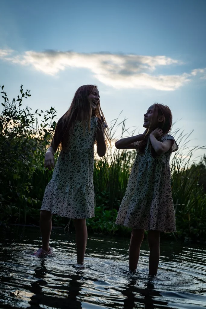 Two girls in matching patterned dresses smiling and playing in a shallow stream at sunset with tall grass and a partly cloudy sky in the background.