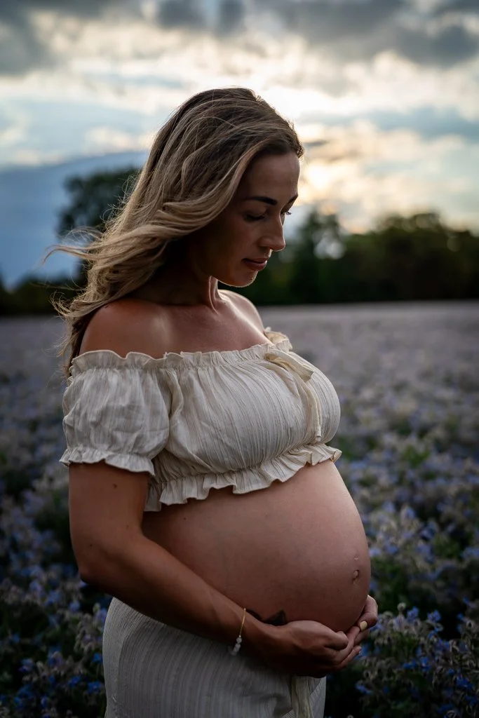 A pregnant woman standing in a field of purple flowers, softly smiling and looking down at her belly during sunset.