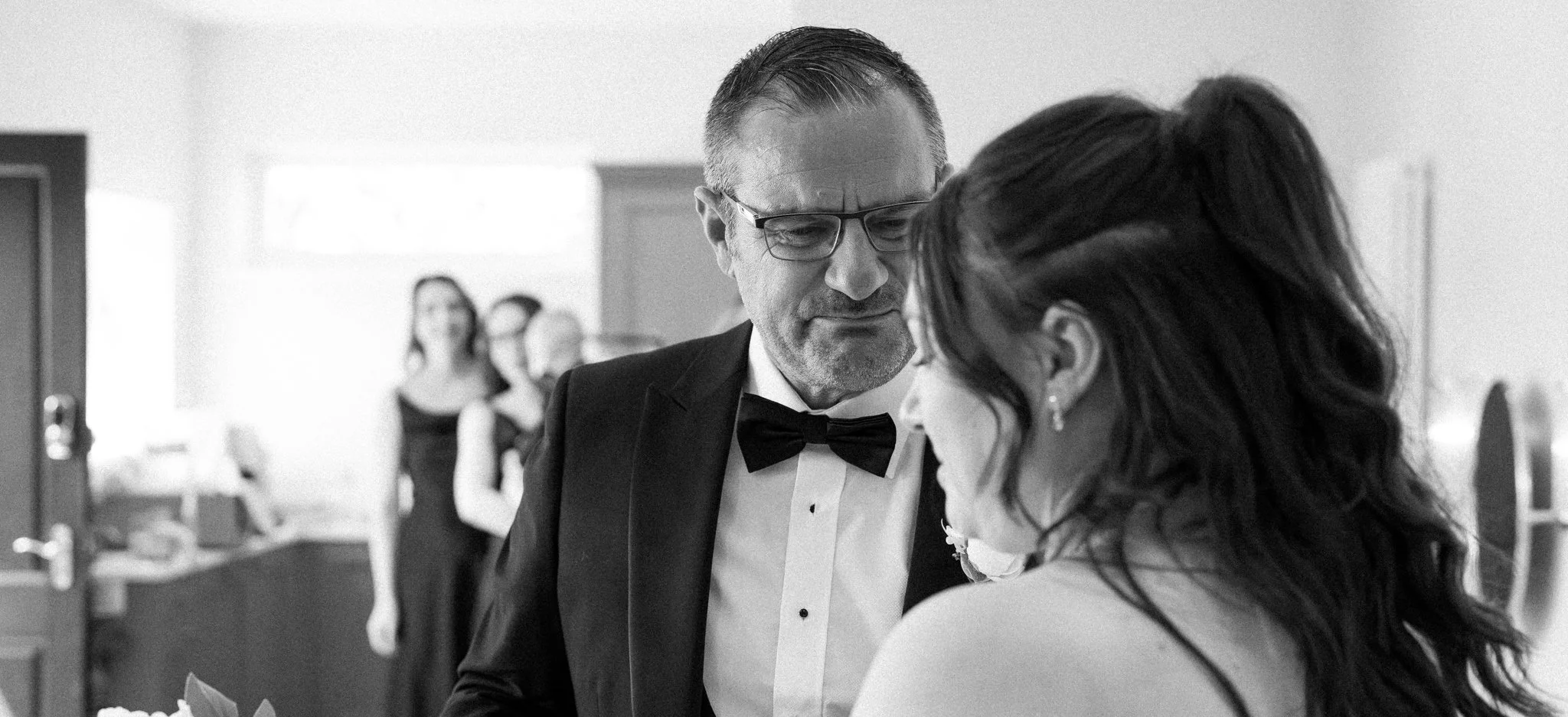 A black-and-white photograph of a man in formal attire with glasses and a woman with long dark hair leaning close to each other, possibly sharing a private moment at a social gathering. Anne Wills Photography Maidens barn Essex