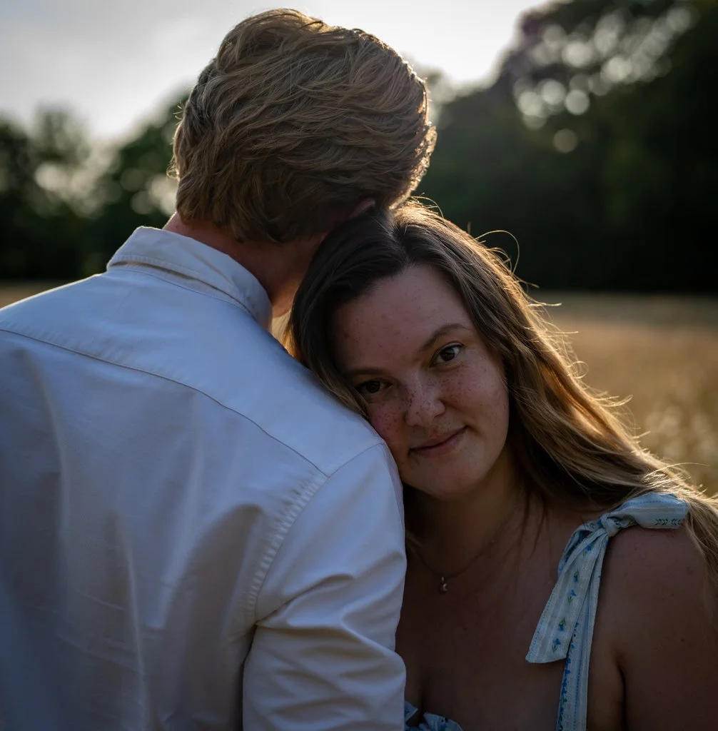 A young woman with long brown hair and freckles leans her head on a man's shoulder. The man has light brown hair and is wearing a light-colored shirt. They are outdoors during sunset in a field with trees in the background. Anne Wills Photography. Br