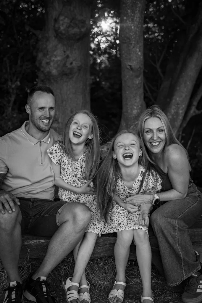 A happy family of four smiling and laughing outdoors in a wooded area, sitting on a log.